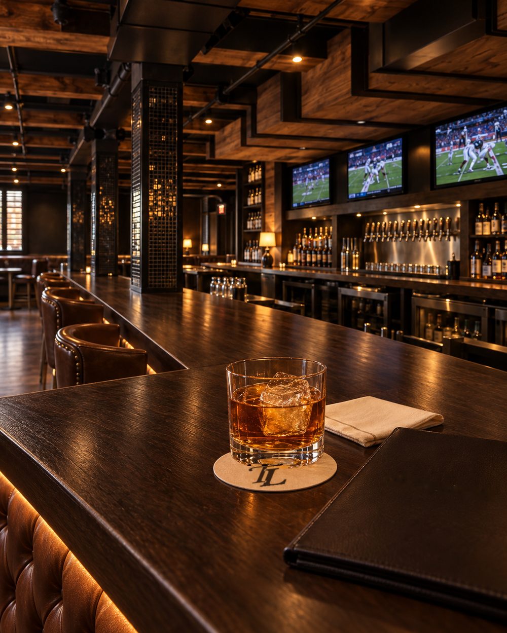 A whiskey over a single large cube on a Letterman coaster at the bar, the back bar and the screens behind it.